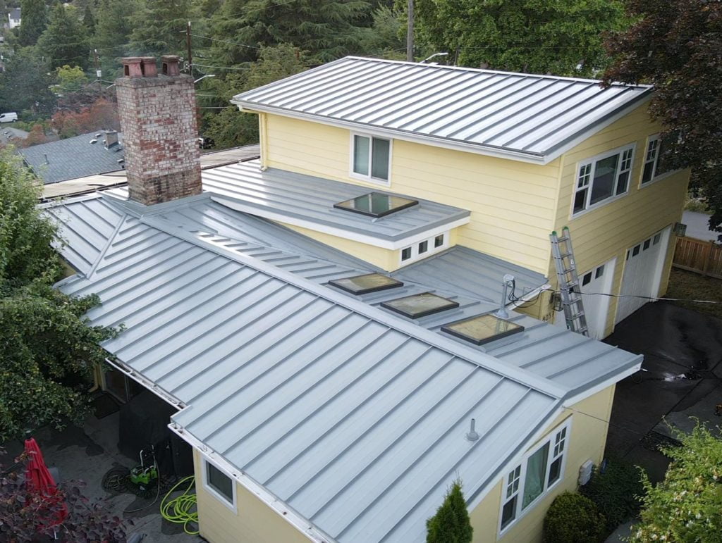 Aerial view of a two-story yellow house with a gray metal roof. The roof, featuring multiple sections with skylights and an attic venting system, also has a chimney and a ladder leaning against the side. The house is surrounded by trees.